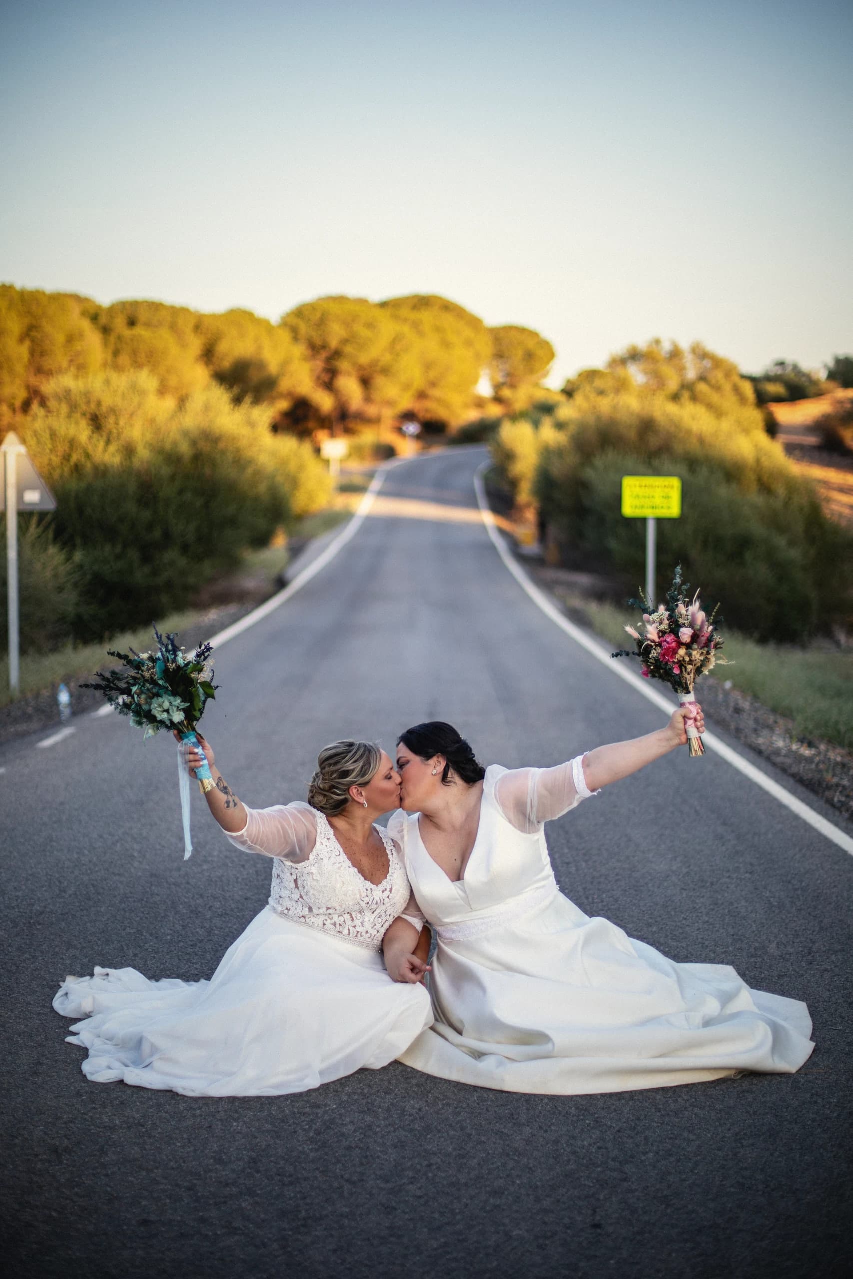 Los novios besándose sentados en mitad de una carretera vacía al atardecer, con la cola del vestido extendida