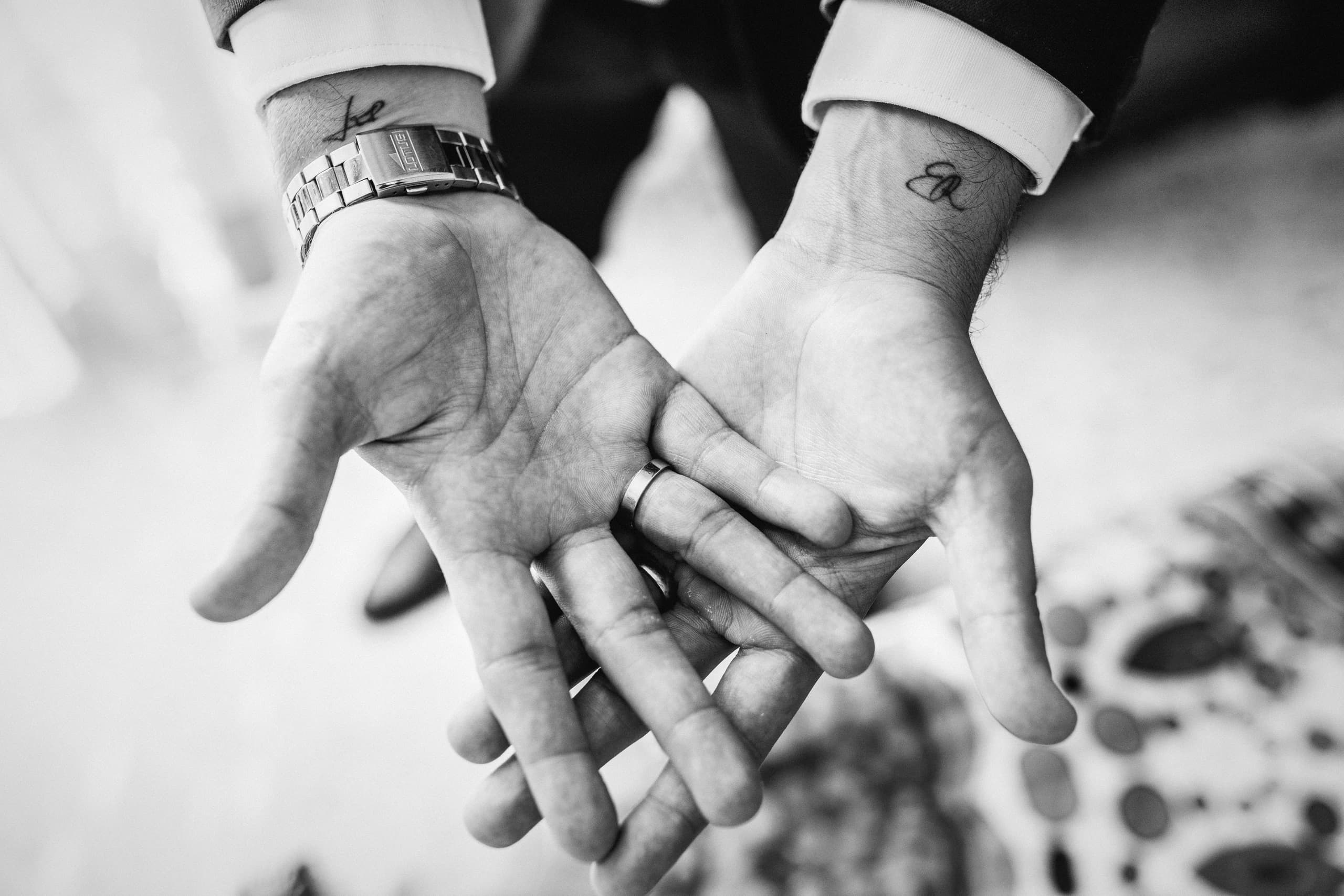 Las manos de los novios abiertas una junto a otra, con las alianzas y los tatuajes de las muñecas, en blanco y negro