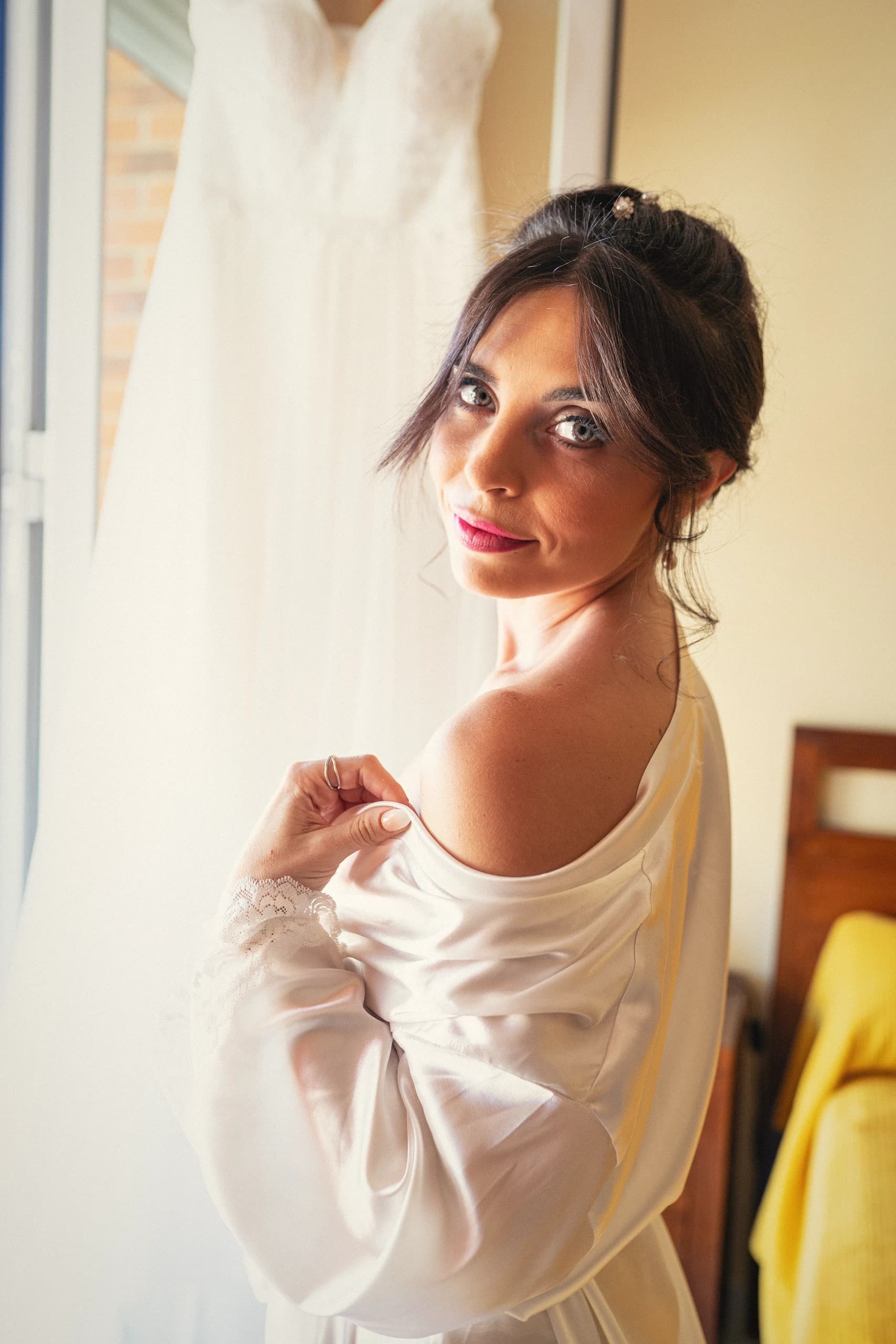 La novia en bata, mirando a cámara junto a la ventana de la habitación mientras se prepara
