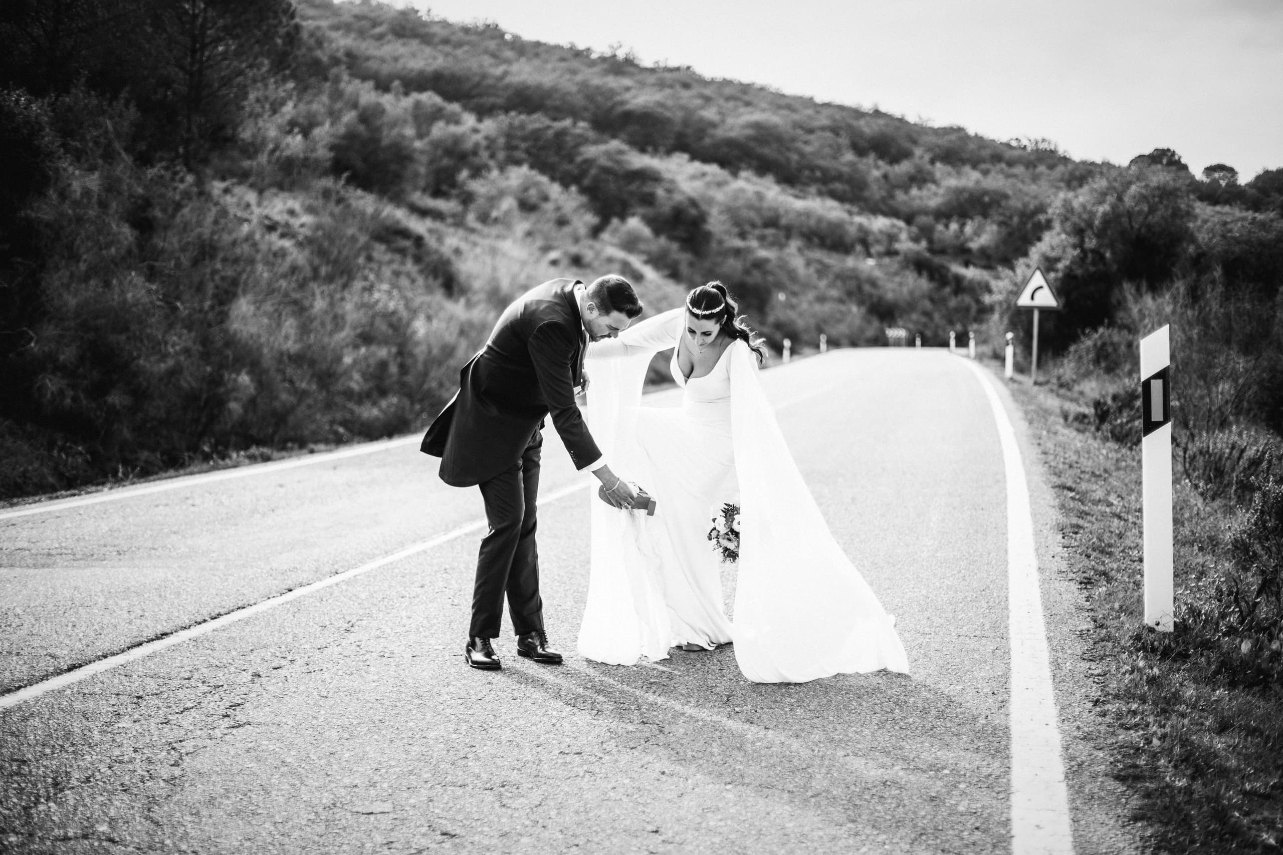 El novio colocando la cola del vestido en la carretera, en blanco y negro