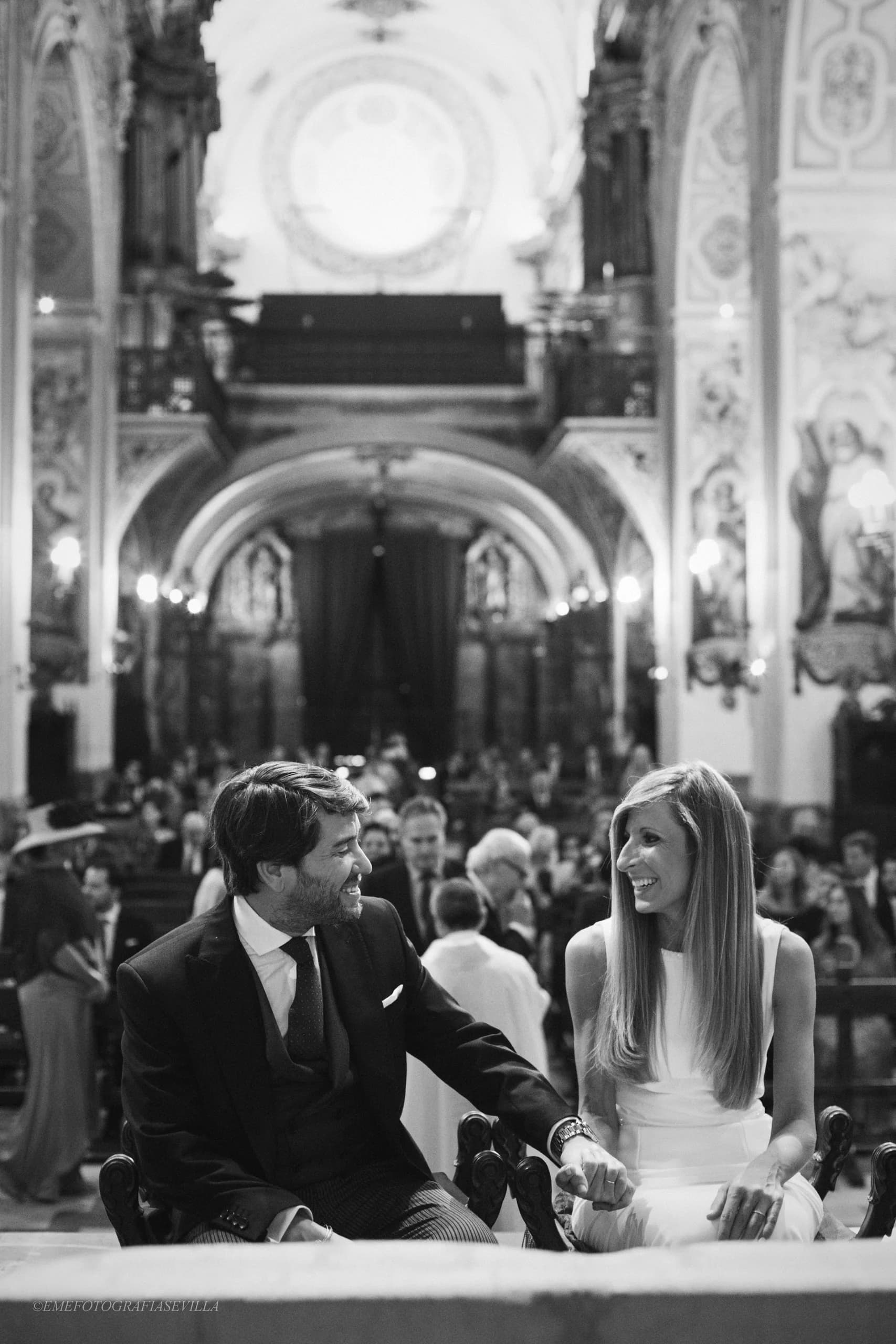 Los novios sentados durante la ceremonia, con la iglesia entera detrás, en blanco y negro