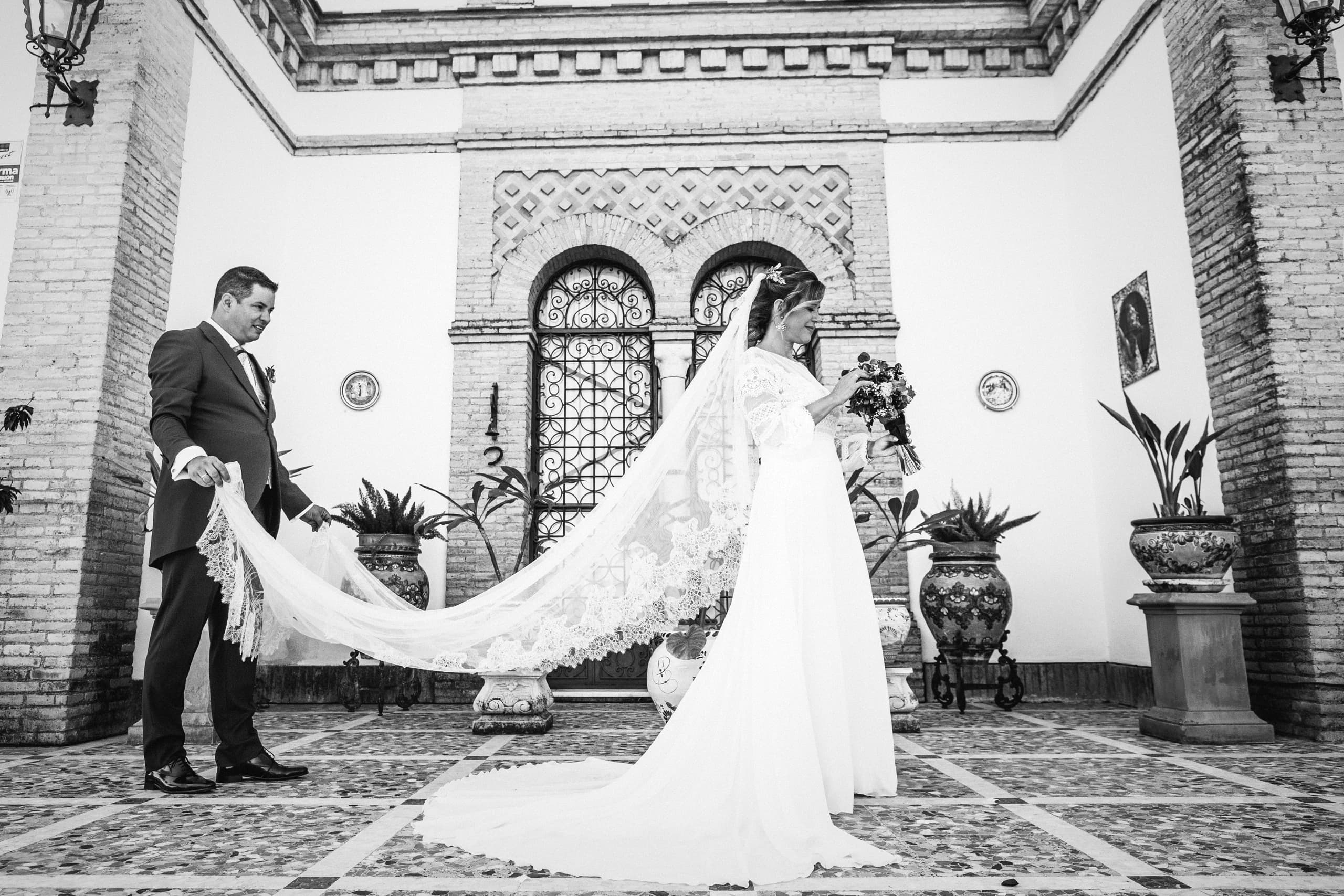 El novio sosteniéndole la cola del vestido en el patio de la hacienda, en blanco y negro