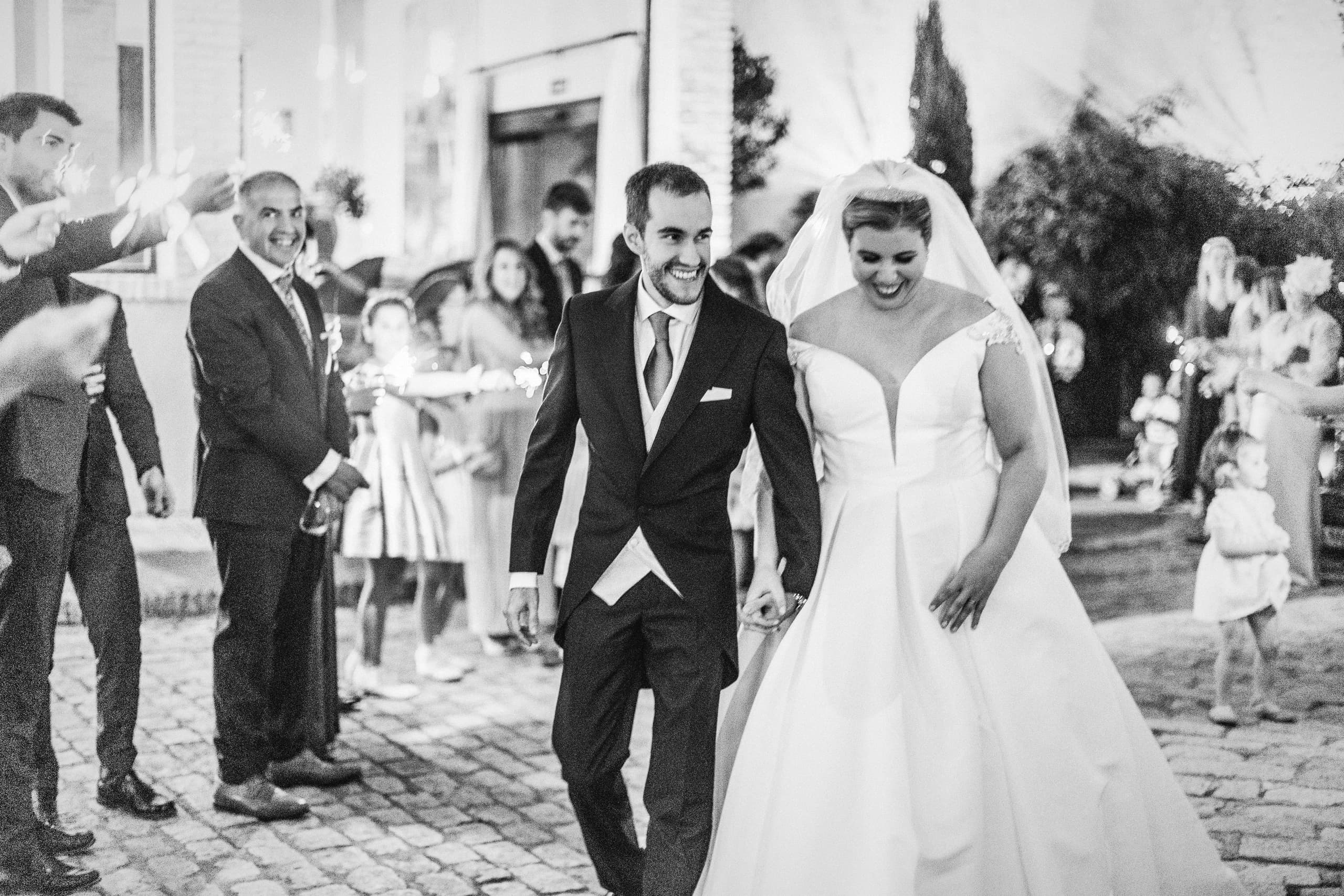 Los novios entre los invitados a la salida, con los brazos en alto, en blanco y negro