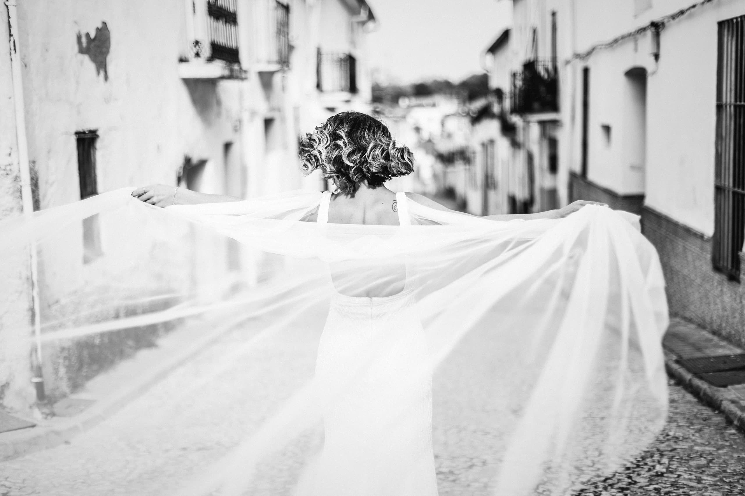 La novia de espaldas con el velo extendido entre las casas blancas, en blanco y negro