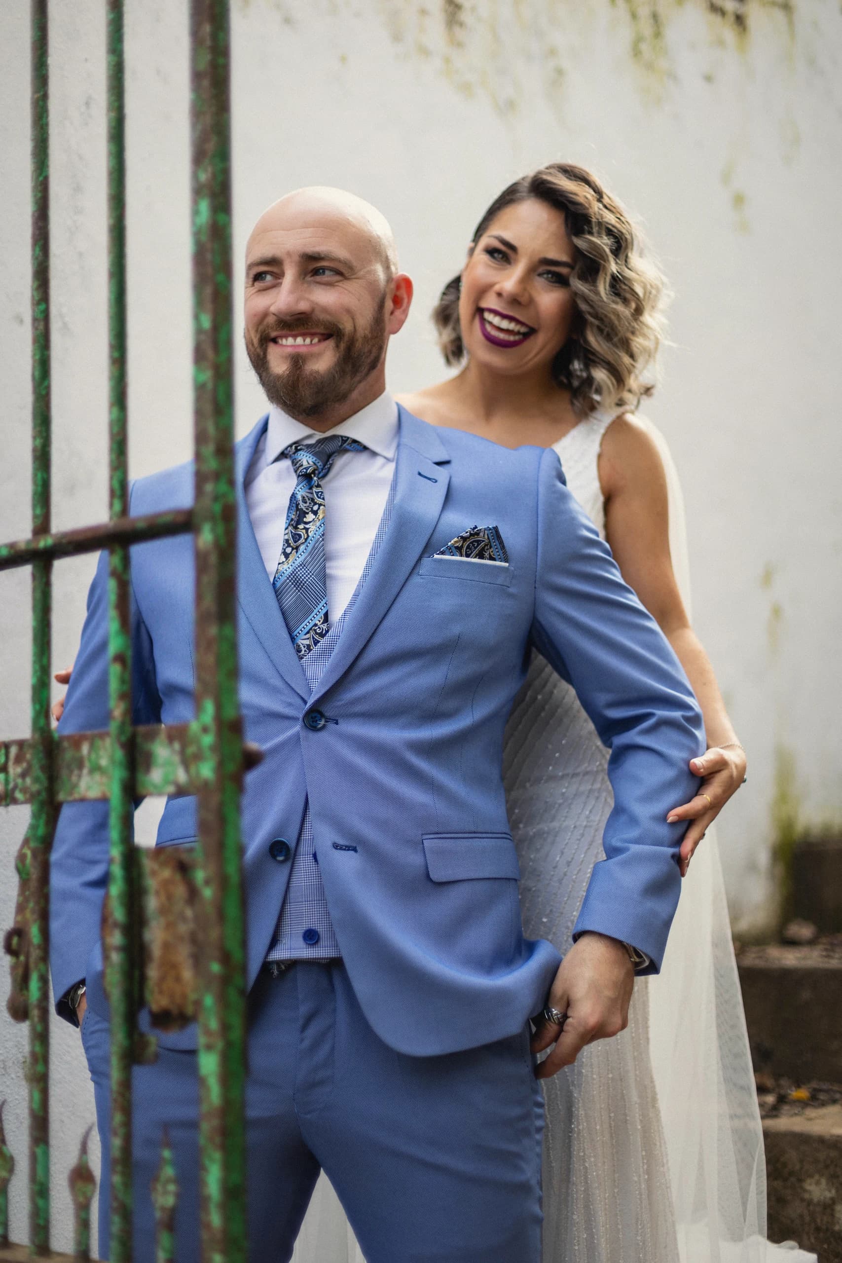 El novio de traje azul y la novia tras una verja verde, sonriendo
