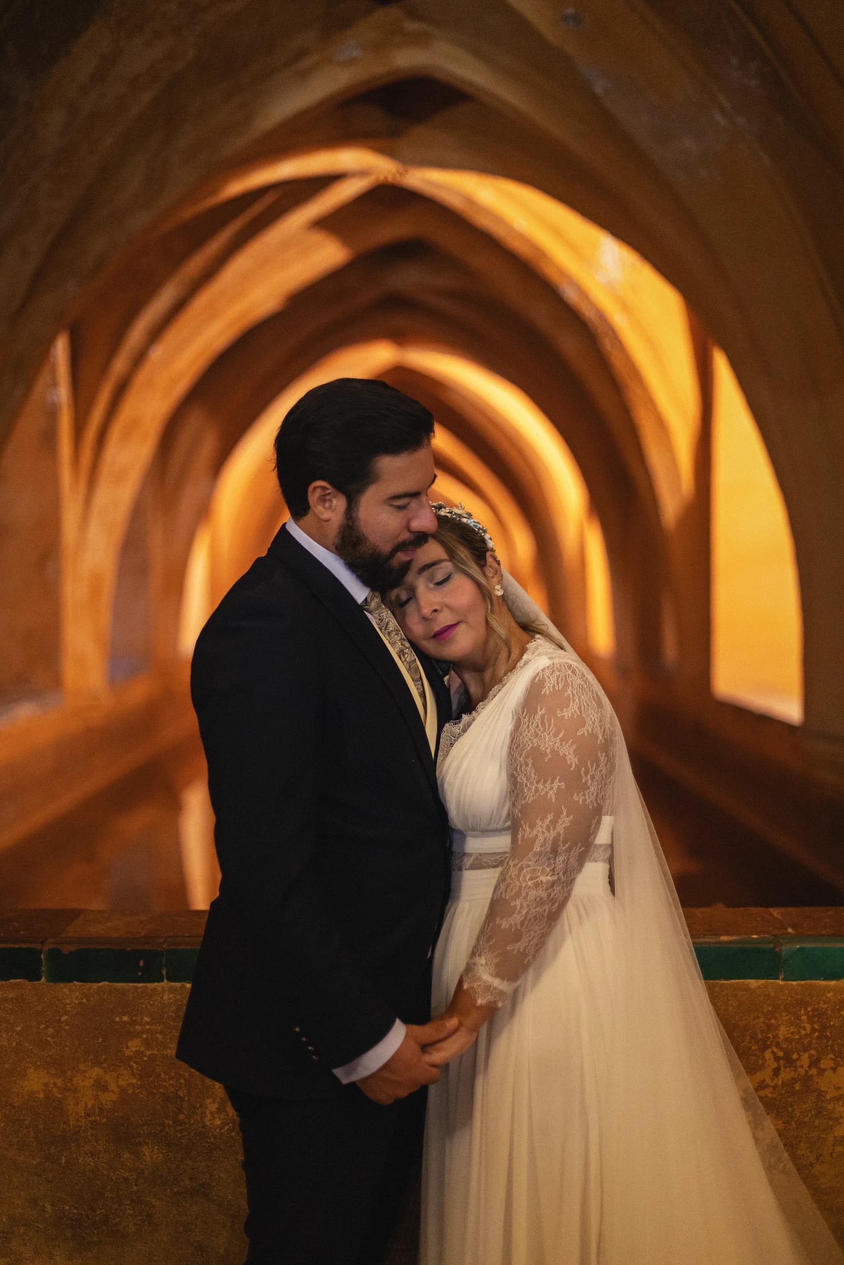 La novia apoyada en el pecho del novio bajo los arcos, a media luz