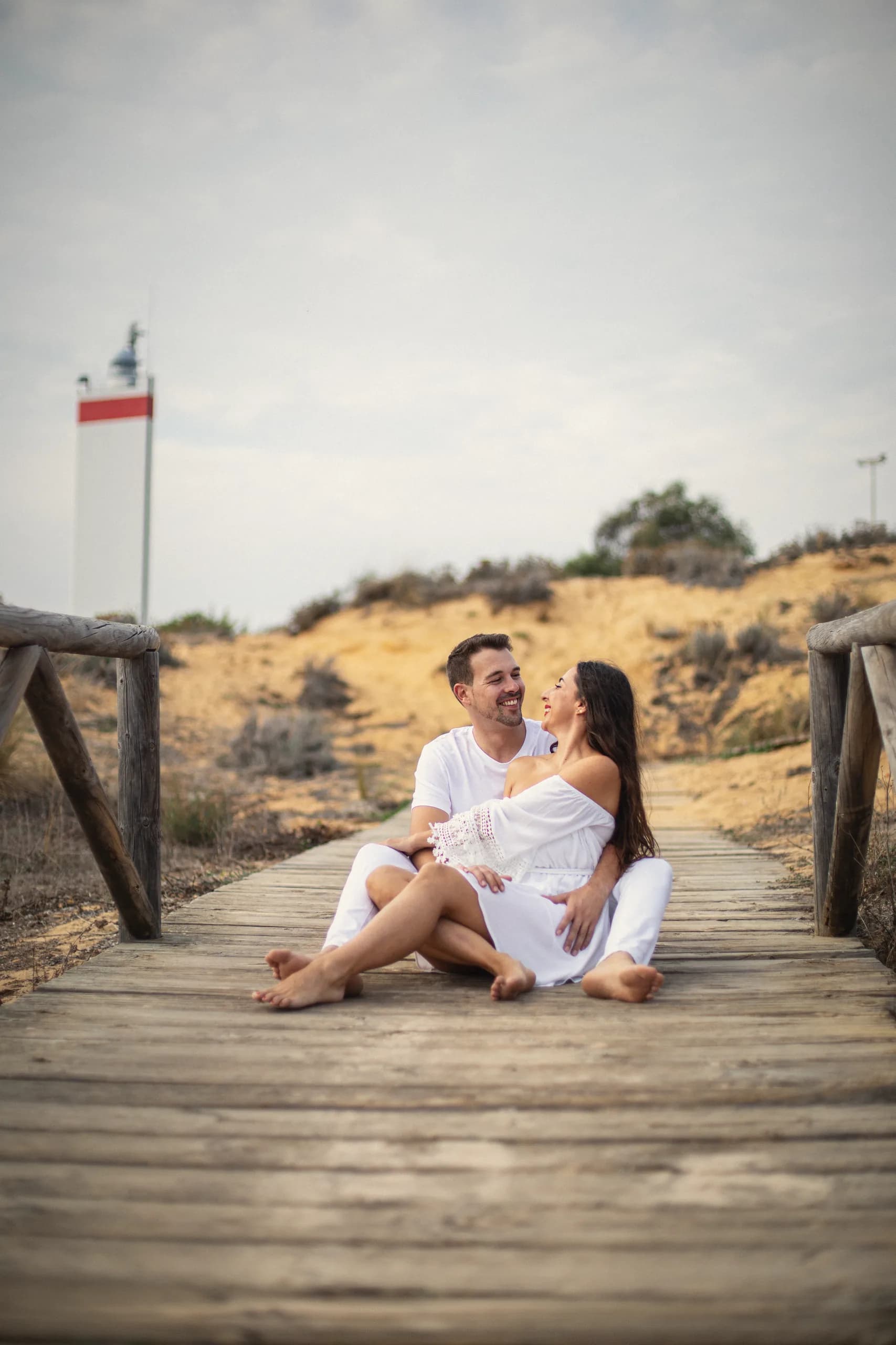 Los dos sentados en la pasarela de madera, riéndose, con el faro al fondo