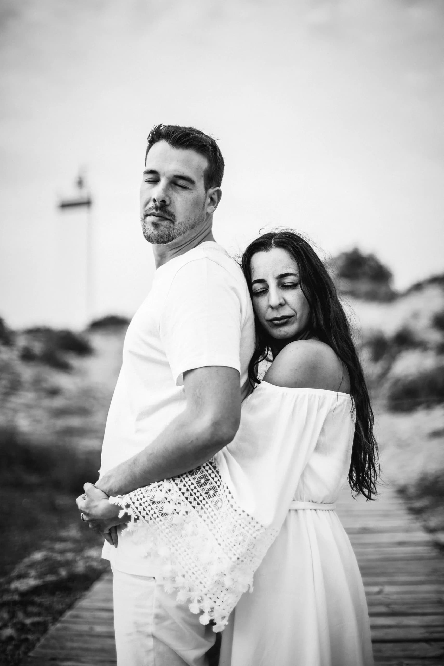 Ella abrazada a su espalda en la pasarela, con el faro detrás, en blanco y negro