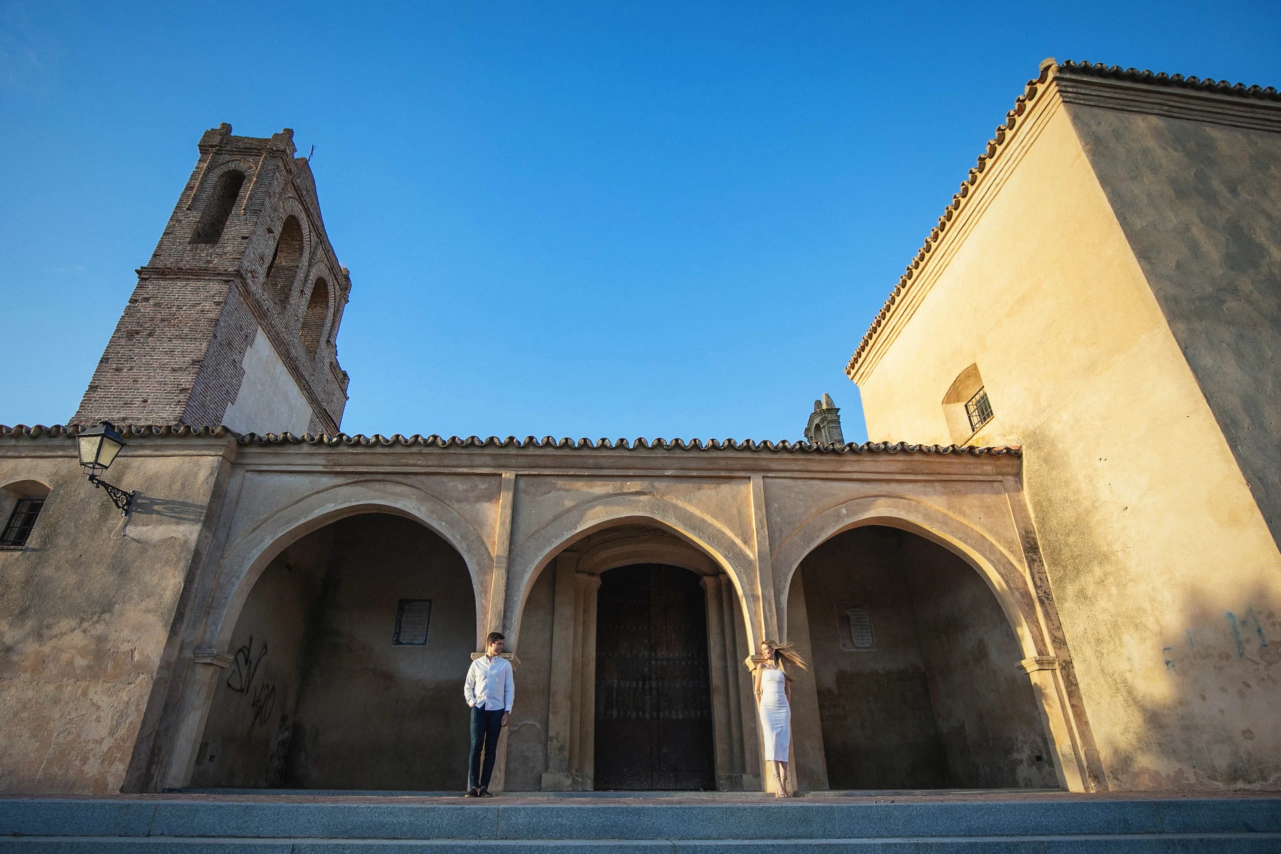 La pareja diminuta bajo los arcos de la iglesia, con la torre al sol
