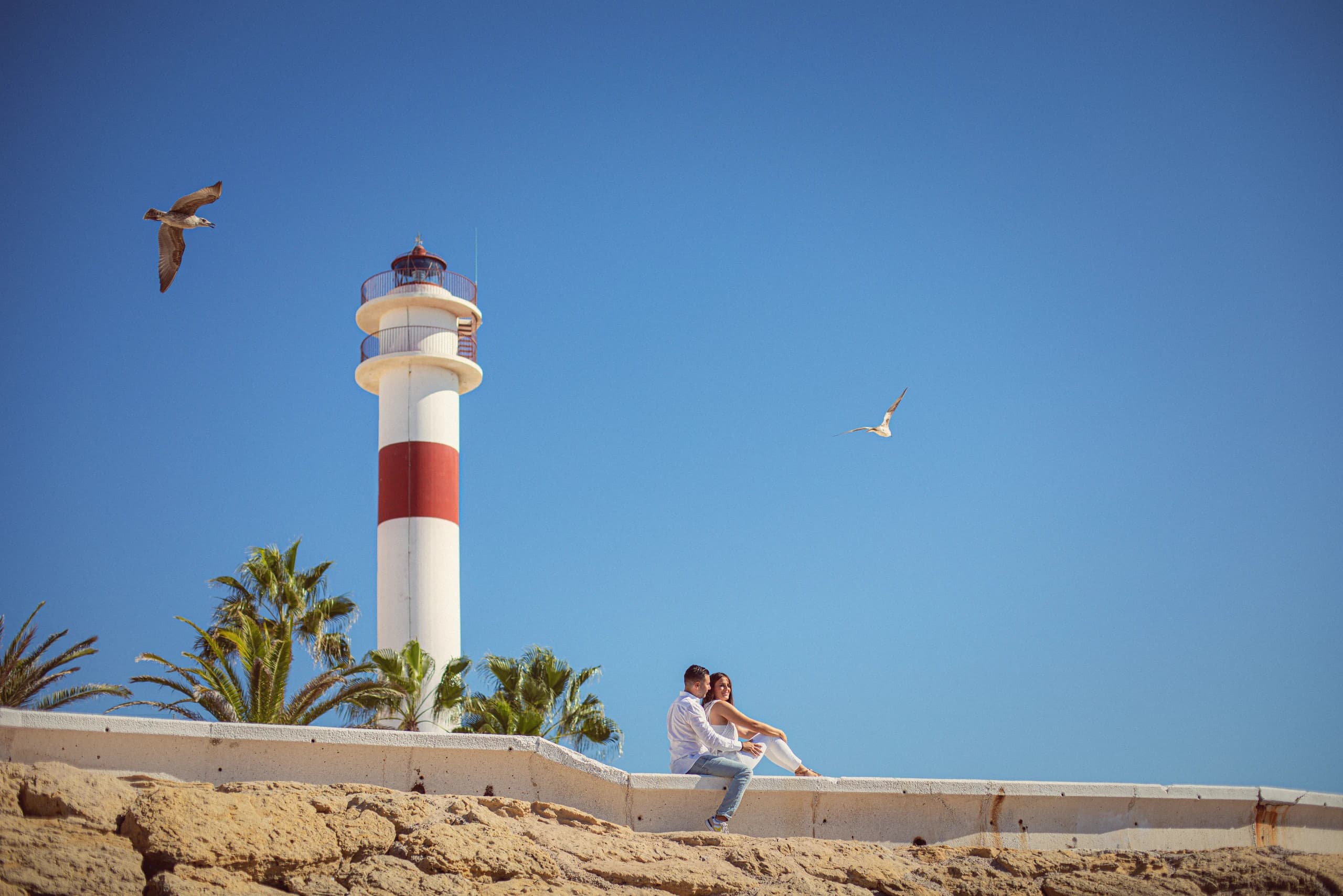 La pareja sentada en el muro con el faro detrás