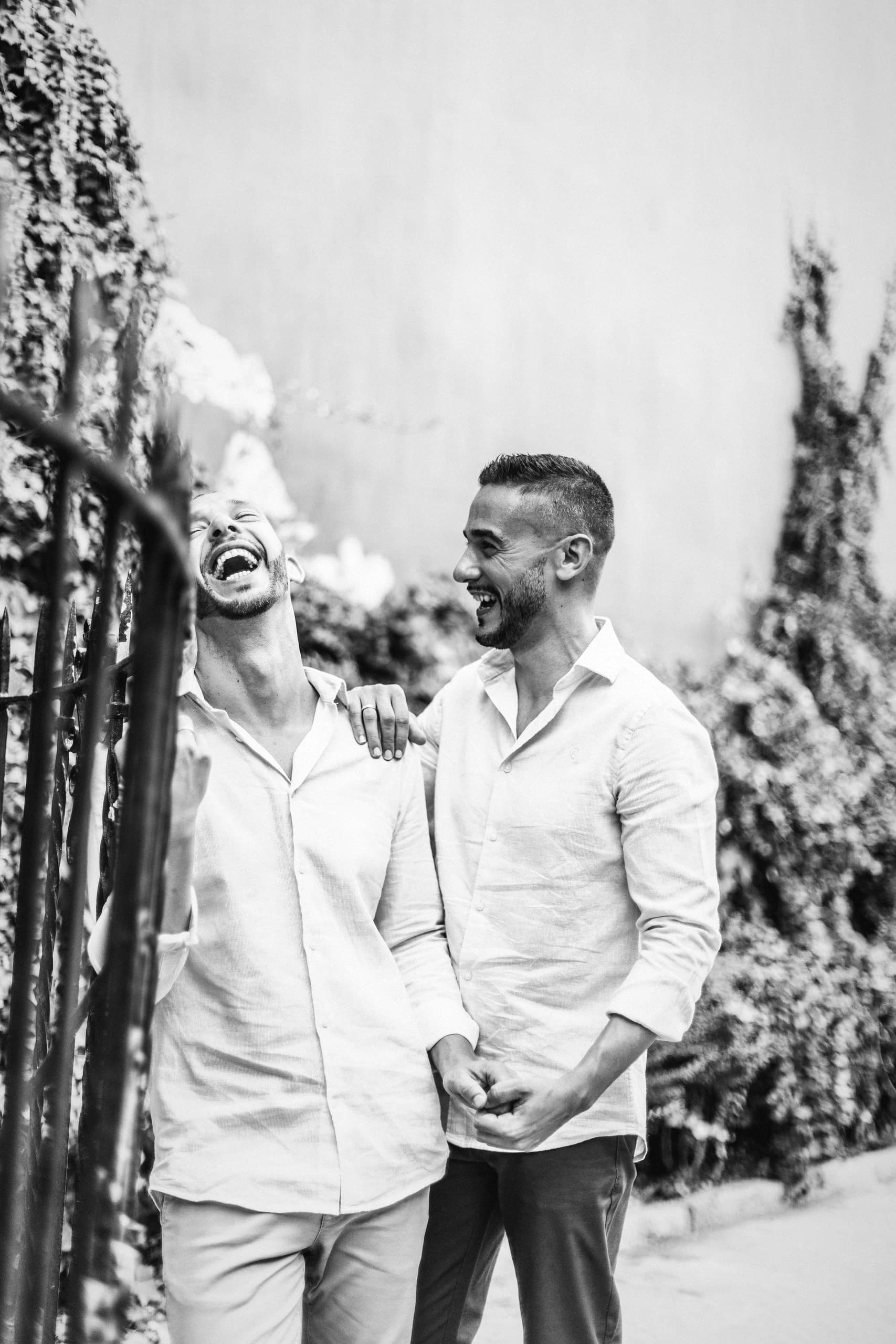 Uno de los dos rompiendo a reír junto a la reja cubierta de plantas, en blanco y negro