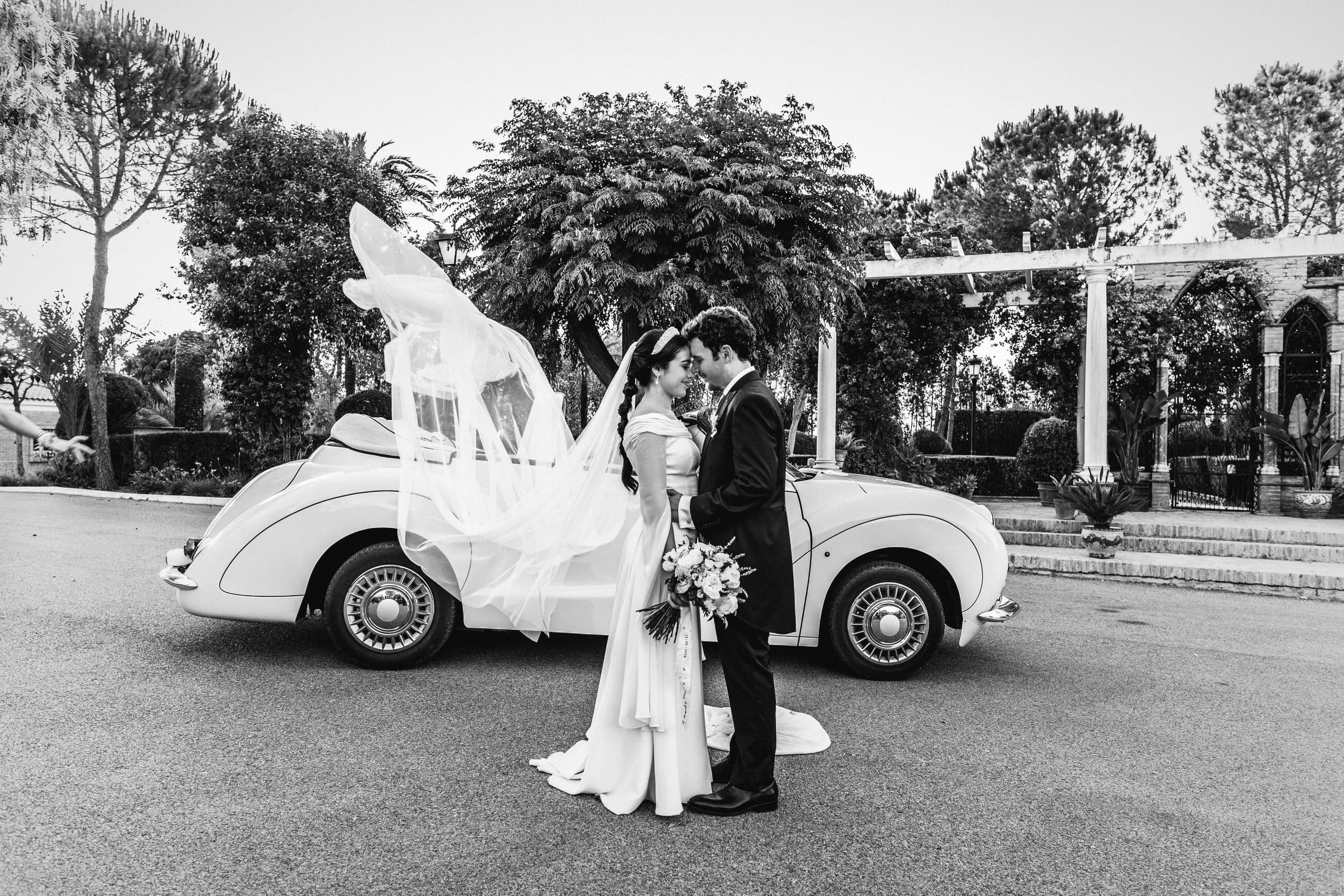 Los novios junto al escarabajo blanco, con el velo al viento, en blanco y negro