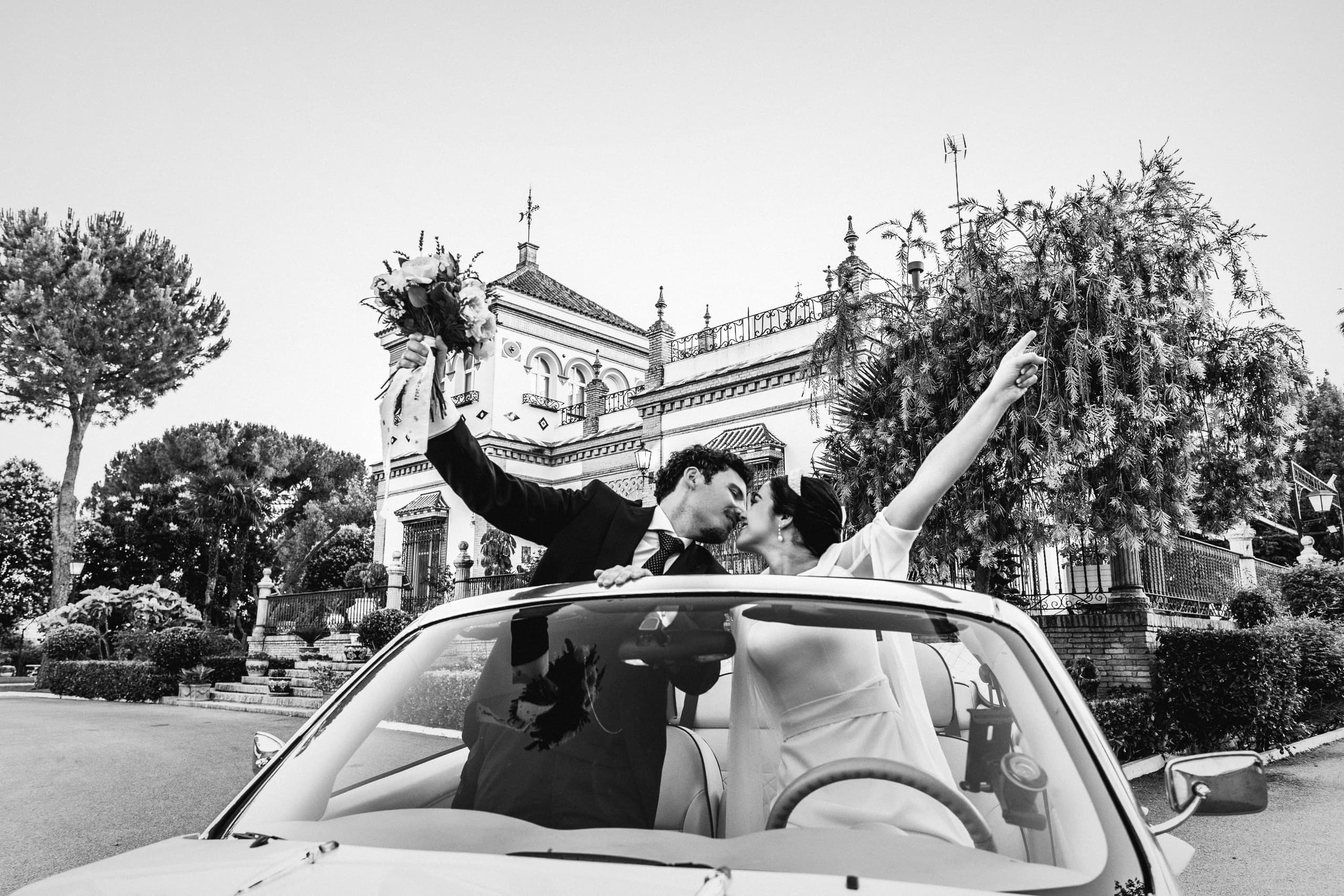 Los novios de pie en el descapotable, con los brazos en alto, en blanco y negro
