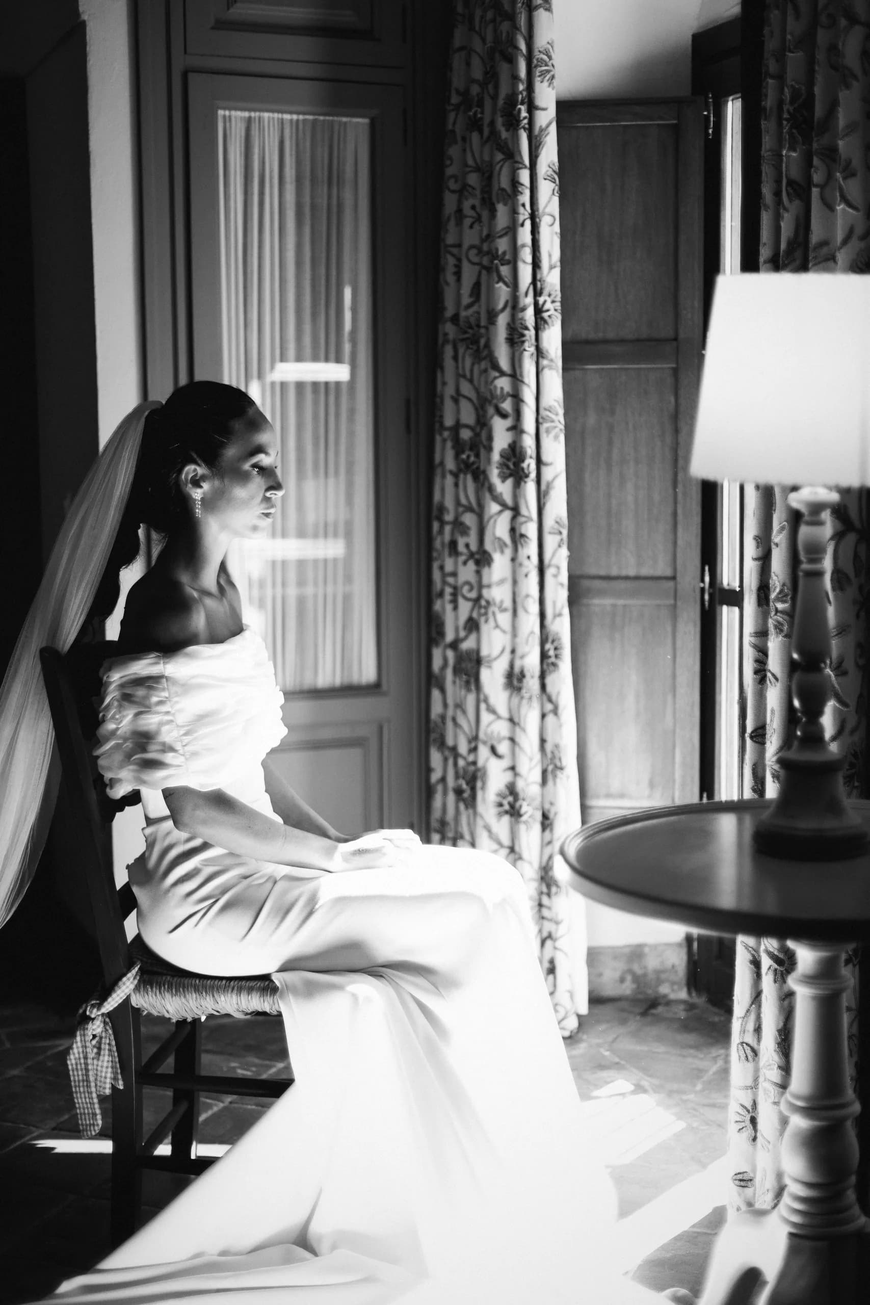 La novia sentada junto a la ventana con el vestido extendido, en blanco y negro