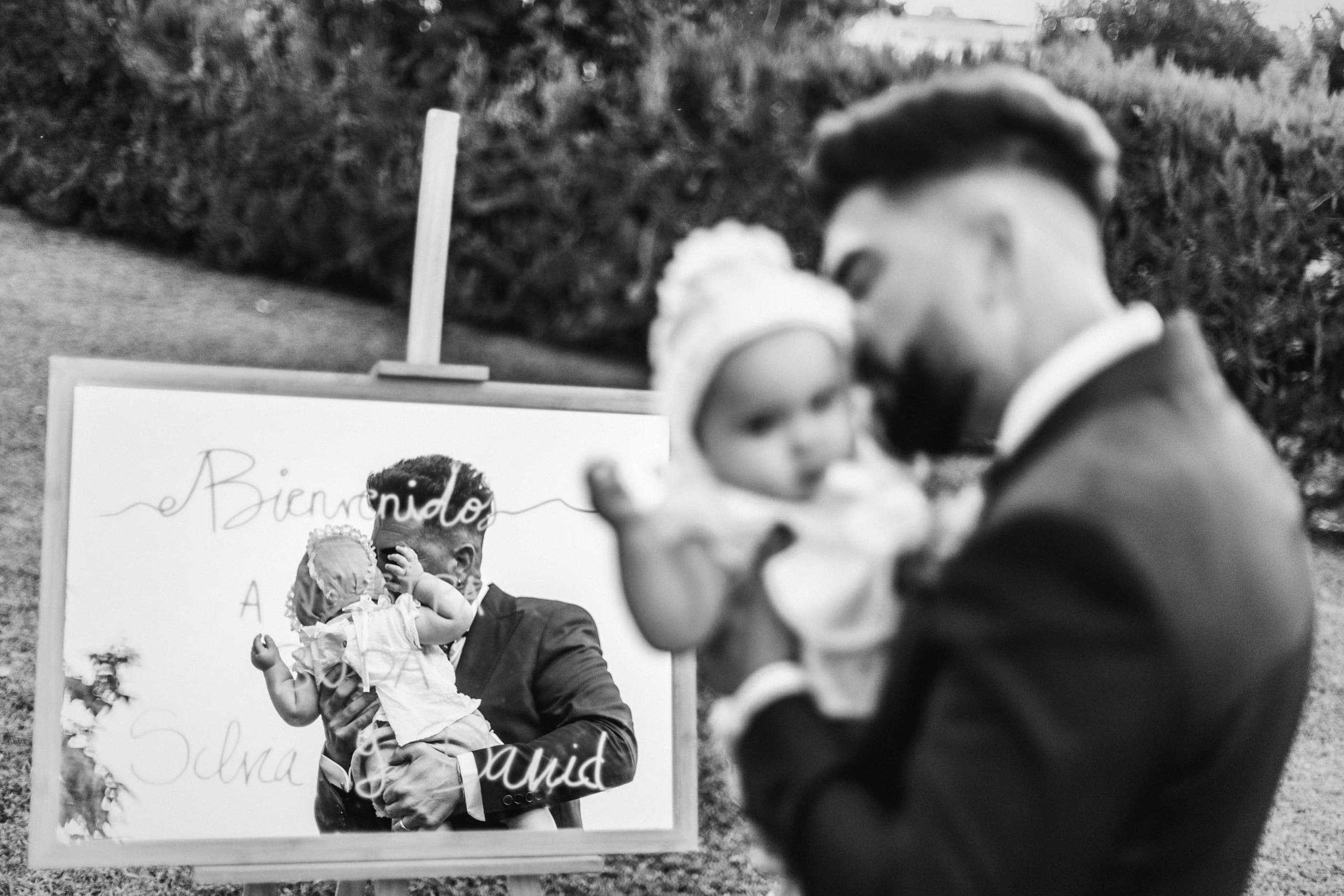 El novio con su hija en brazos junto al cartel de bienvenida, en blanco y negro