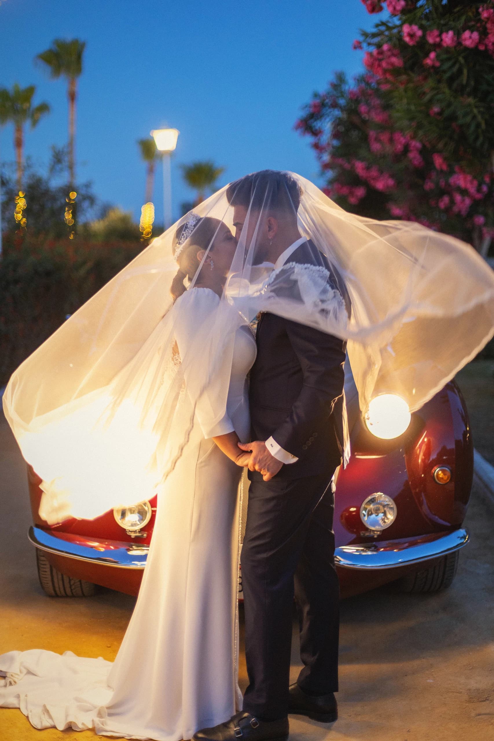 Los novios besándose delante del coche clásico, con el velo al viento y el sol bajo detrás