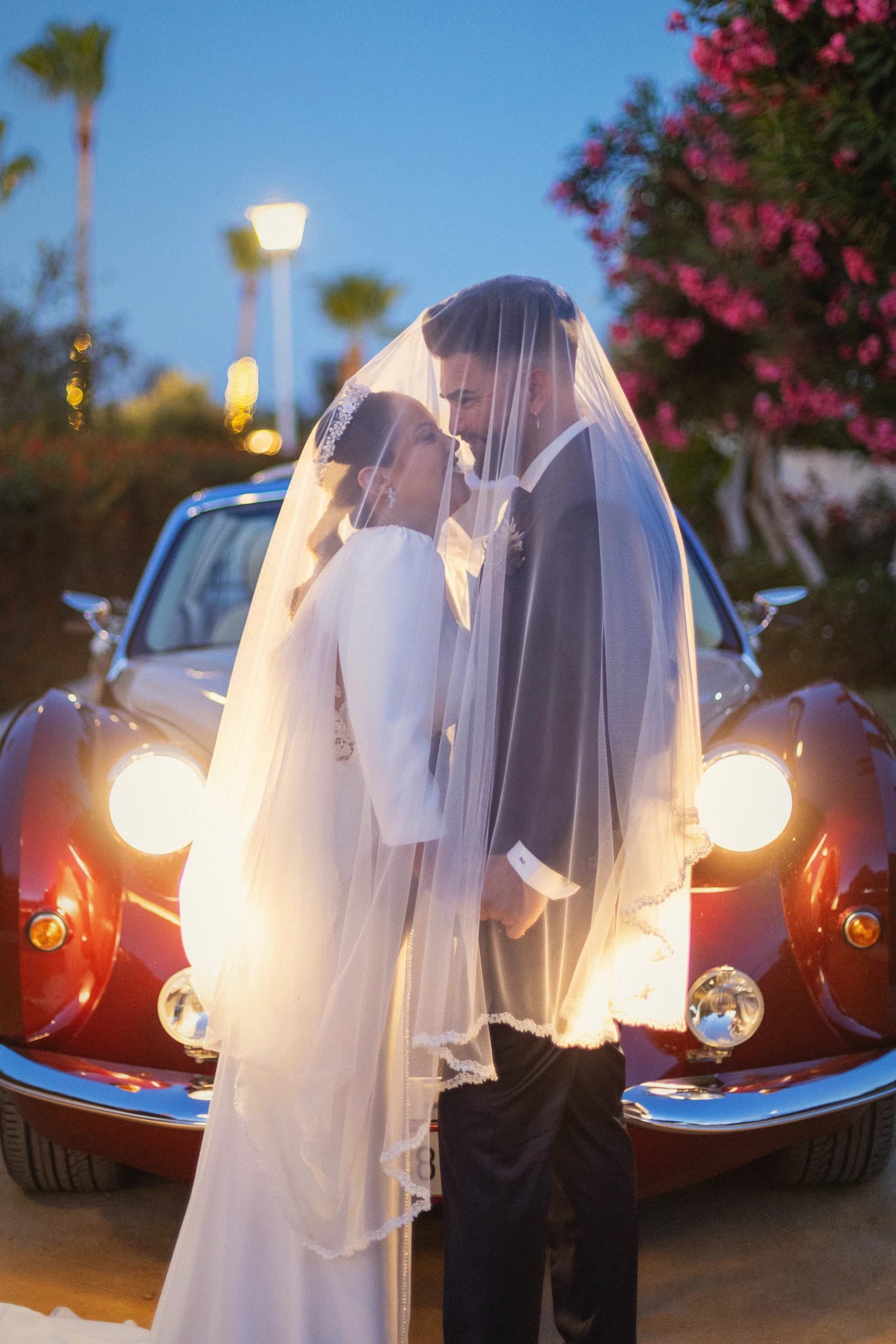 La novia con el velo levantado ante los faros encendidos del coche, al anochecer