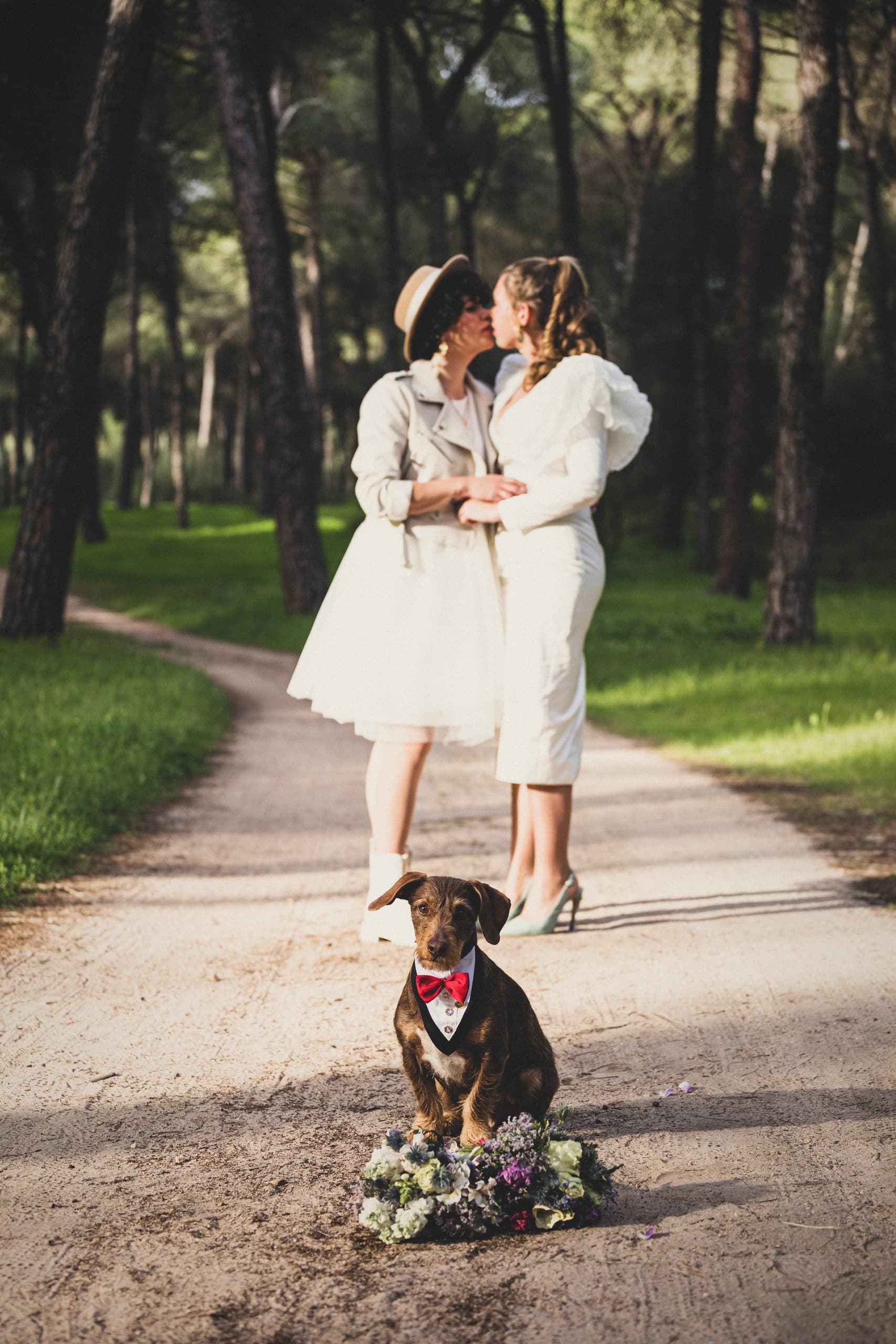 Las dos novias besándose al final del camino del pinar, con el perro sentado en primer plano con pajarita y una corona de flores